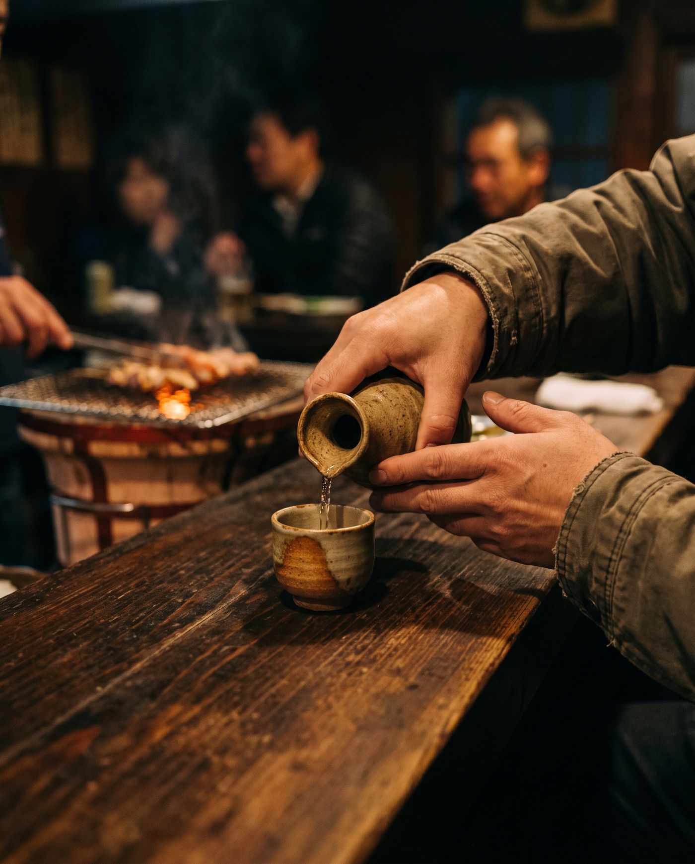 カウンターで、徳利からおちょこへ冷酒を注ぐ手元。奥で串が焼かれている